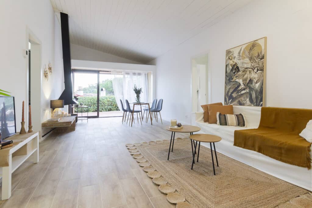 Una sala de estar moderna con un sofá blanco, mesas de café de madera y un comedor, perfecta para la puesta en escena de una casa. Las grandes ventanas ofrecen vistas a una exuberante vegetación, que complementa el piso de madera clara. Una pieza de arte decorativo en la pared le agrega carácter a este acogedor espacio listo para alquilar.