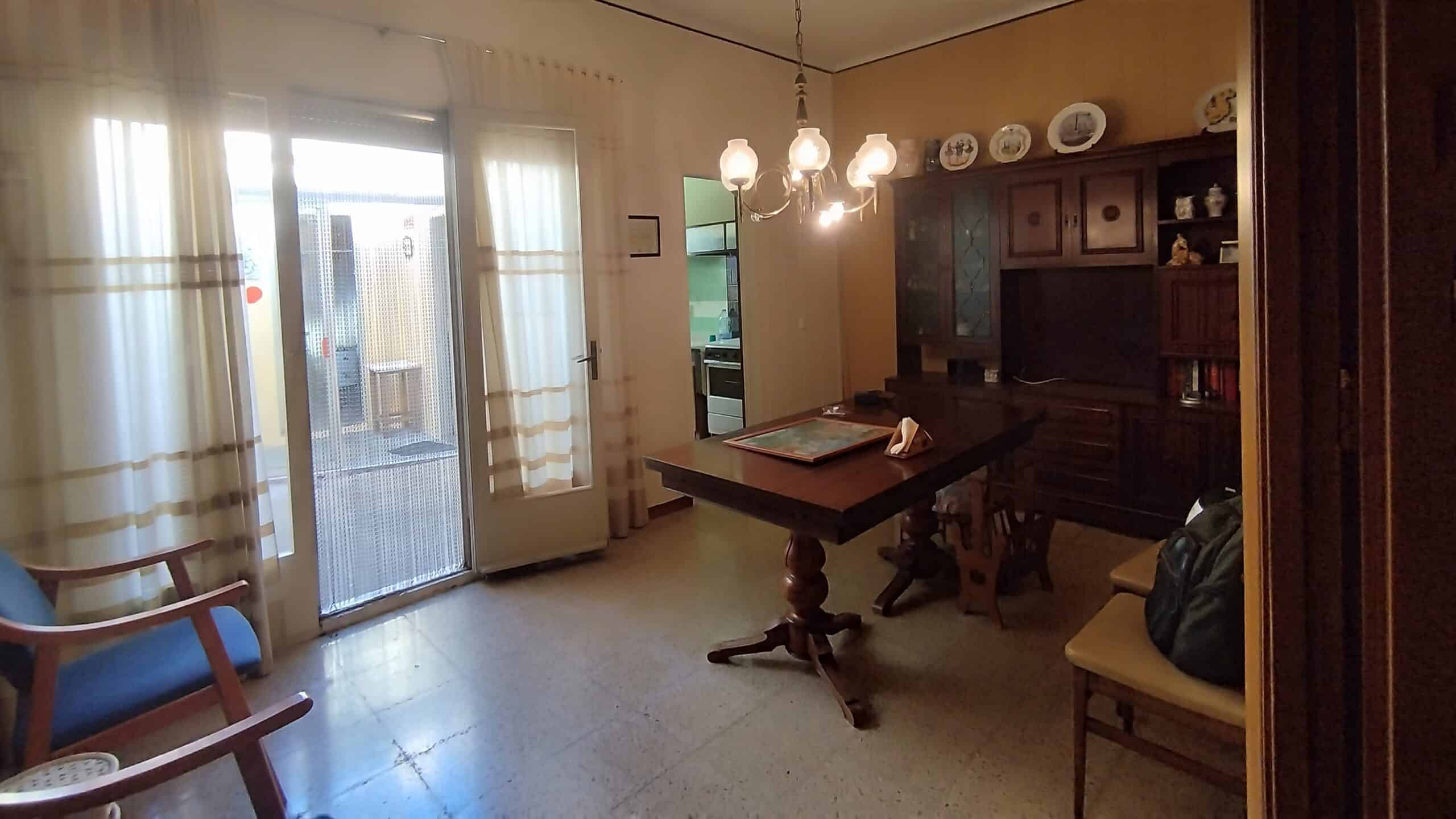 Un comedor con una mesa de madera, sillas, lámpara de araña y un armario. Las puertas francesas conducen a un área al aire libre y se puede ver una cocina adyacente a través de una puerta.