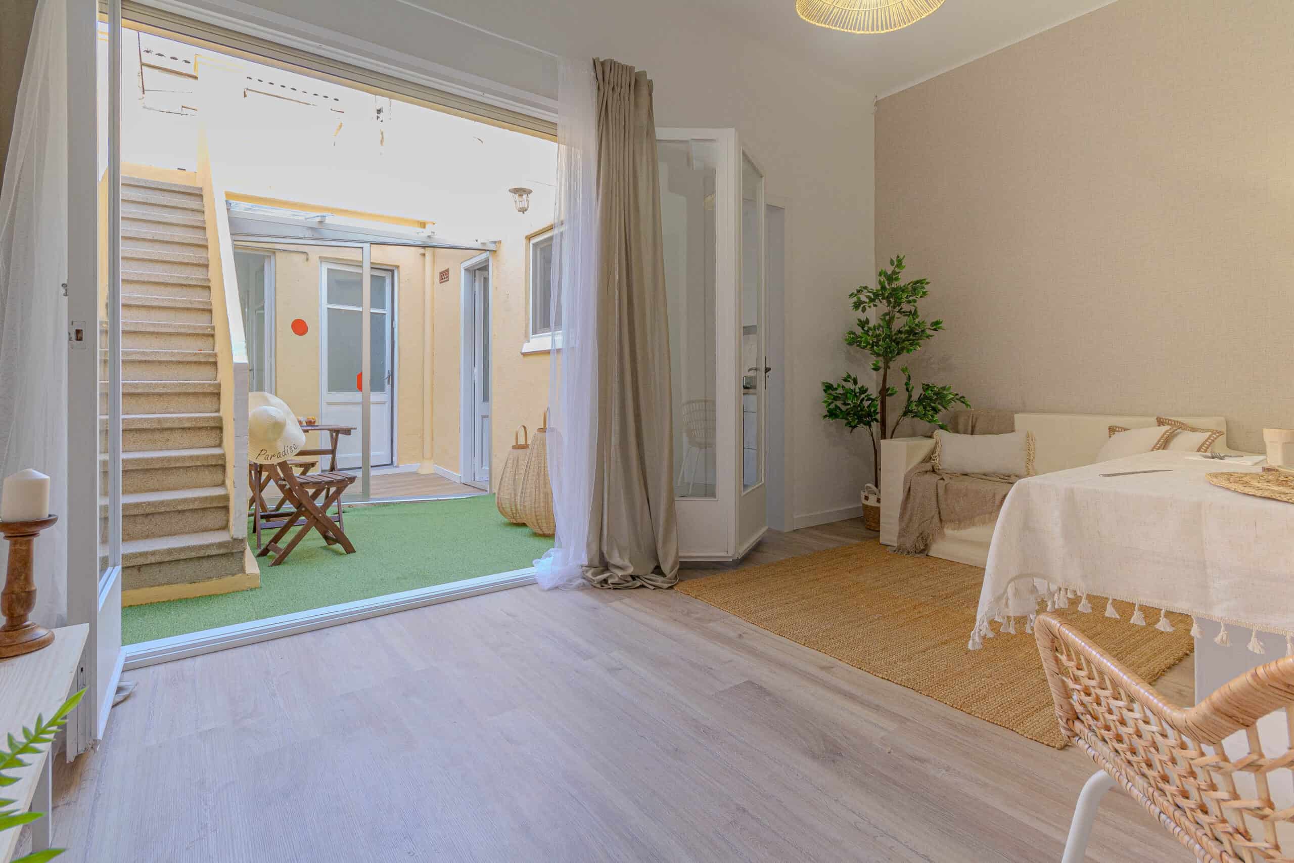 Espacio interior luminoso con suelo de madera, una mesa pequeña y un sofá. Una puerta abierta revela una escalera que conduce a un área al aire libre con suelo verde y muebles de patio.