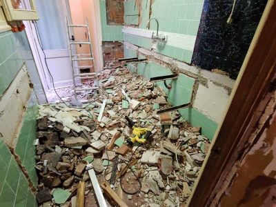 Una habitación estrecha en renovación, con escombros y materiales de construcción esparcidos por el suelo, paredes parcialmente revestidas y una escalera cerca de una puerta.