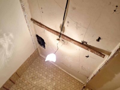 Habitación en proceso de renovación con cableado a la vista y techo sin pintar. Una única bombilla cuelga, iluminando el papel pintado estampado y las paredes parcialmente decapadas.