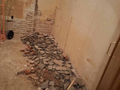Una habitación en renovación con azulejos rotos y escombros en el suelo, ladrillos expuestos en una pared y una carretilla de mano en la esquina.