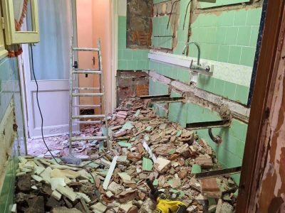 Una habitación parcialmente demolida con escombros en el suelo, paredes de ladrillo a la vista, baldosas verdes y herramientas de construcción, incluido un taladro y una escalera.