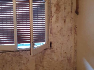 Una habitación con papel tapiz descascarado, una ventana abierta y persianas de madera. El suelo está cubierto de trozos de papel tapiz rotos.