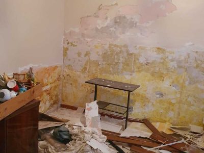 Habitación en proceso de renovación con pintura descascarada y yeso a la vista en las paredes, escombros y trozos de madera rotos esparcidos por el suelo. En la esquina hay un pequeño estante.