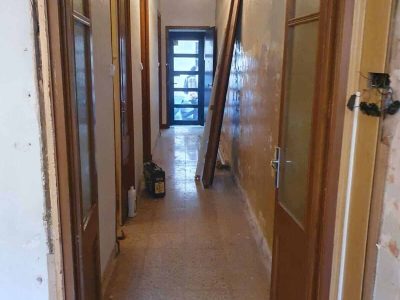 Pasillo estrecho con puertas a ambos lados, yeso sin terminar en la pared derecha y materiales de construcción esparcidos. Una puerta entreabierta al final conduce a una habitación con poca luz.