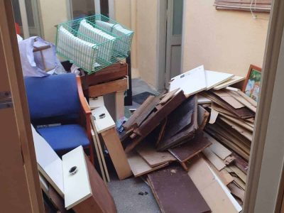 Una habitación desordenada con tablas de madera, una silla azul, una pequeña jaula verde y objetos diversos esparcidos por el suelo.