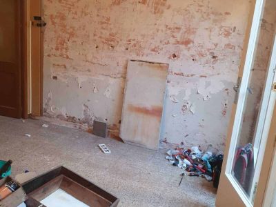 Una habitación con pintura descascarada en las paredes, escombros esparcidos por el suelo y una puerta abierta. Un armario retirado yace de lado con una rejilla de alambre cerca.