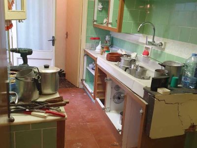 Una cocina pequeña y desordenada con paredes de azulejos verdes. Hay ollas, utensilios de cocina y una botella de agua esparcidos. El armario inferior está roto y deja al descubierto las tuberías. Un reloj marca las 10:10.