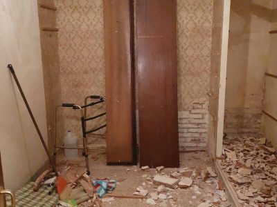 Una pequeña habitación en proceso de renovación con papel tapiz, escombros en el piso y dos paneles de madera apoyados contra la pared. Se ven una serie de estantes y algunas herramientas dispersas.