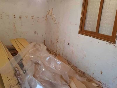Una habitación parcialmente renovada con paredes desnudas, una ventana, una lámpara de araña y tablones de madera y láminas de plástico dispersos en el suelo.