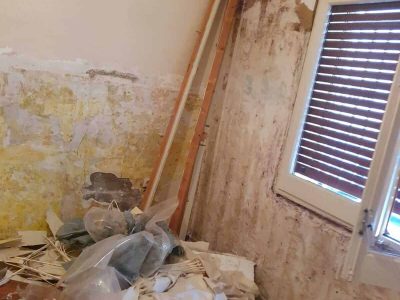 Una habitación con pintura descascarada, moho en las paredes y escombros, incluidas vigas de madera y bolsas de plástico, esparcidos por el piso. Una ventana con persianas de madera está parcialmente abierta.