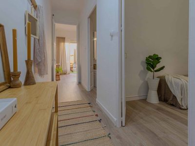 Vista de un pasillo con suelo de madera, una mesa con elementos decorativos a la izquierda y una planta en maceta blanca a la derecha. Al fondo se ve una habitación con cama y cortinas.