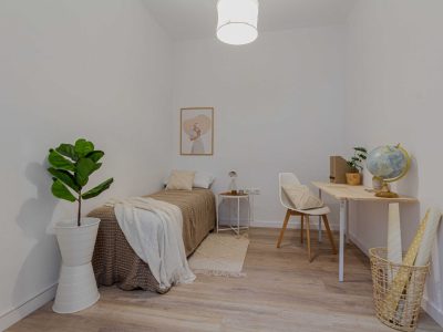 Dormitorio minimalista con cama, escritorio blanco, silla, maceta, globo terráqueo y cuadro mural. Una cesta tejida y una lámpara completan la decoración.