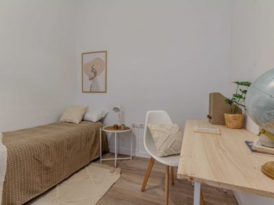 Un dormitorio minimalista con una cama, un pequeño escritorio, una silla y un globo terráqueo. Una obra de arte enmarcada se encuentra en la pared sobre la cama. Los colores neutros dominan la decoración.