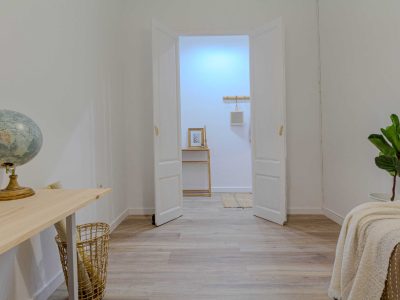 Habitación con suelo de madera, un escritorio con un globo terráqueo, una puerta abierta que da a un pasillo y una planta al lado de una cama cubierta con una manta.