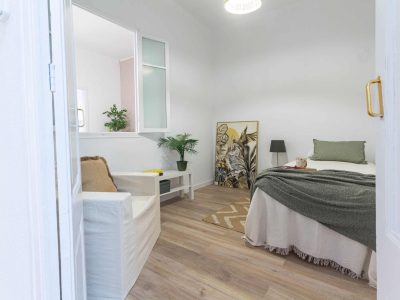 Un dormitorio pequeño y minimalista con una cama, un sillón, plantas y una obra de arte enmarcada en el suelo. Las paredes son blancas y el suelo de madera. Una ventana refleja la luz natural.