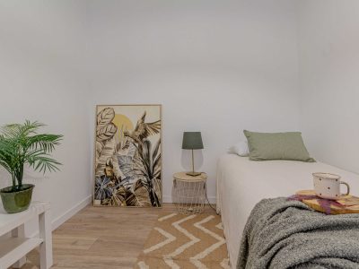 Dormitorio minimalista con una cama individual, una almohada verde, una mesa auxiliar con lámpara, una planta en maceta, una obra de arte enmarcada y una taza sobre una manta. Los tonos neutros crean una atmósfera tranquila.