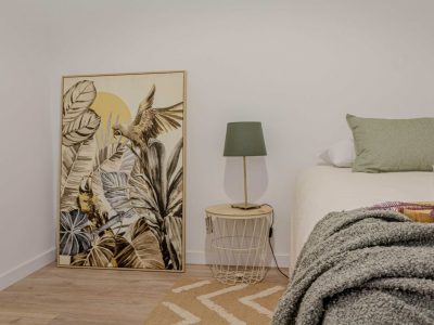 Interior de un dormitorio con un gran estampado botánico apoyado contra la pared, junto a una mesita de noche redonda pequeña con una lámpara. A la derecha, una cama con almohadas verdes y una manta con textura.