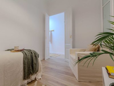 Una habitación acogedora con una cama, un sillón blanco y una pequeña planta. La puerta abierta revela un espacio contiguo luminoso.