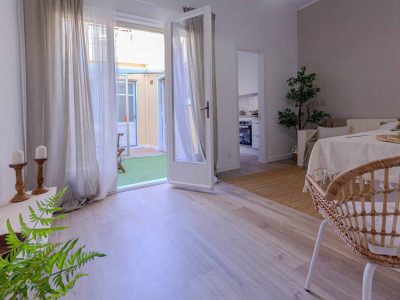Una habitación luminosa y espaciosa con una puerta abierta que da al exterior. El interior cuenta con suelos de madera clara, una mesa con un mantel beige, un pequeño sofá, plantas y cortinas transparentes.